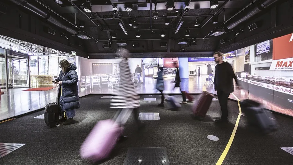 Several people are standing in a room with images from an airport projected on the walls. They have coats and suitcases. There is a yellow strip of tape on the floor. There are lots of technical equipment in the ceiling.