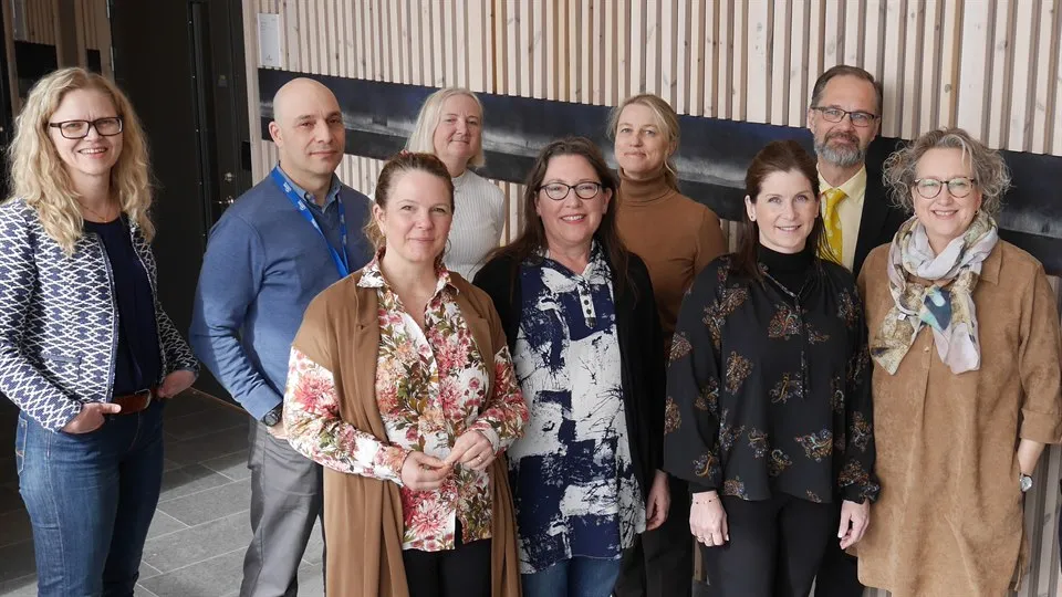 Nio män och kvinnor står tillsammans framför en trävägg. De ser glada ut och tittar in i kameran. 