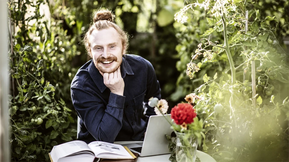 Male student sitting in greenhouse studying with laptop and books.