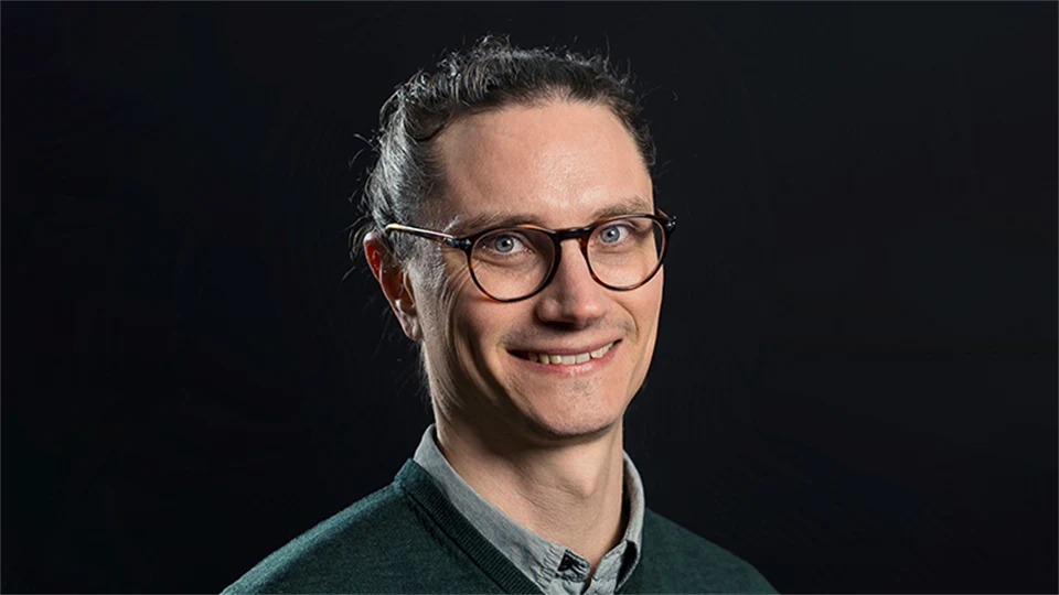 En man i glasögon och mörkt hår ser in i kameran och ler. 