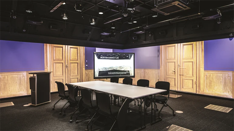 A large room with black floor and ceiling. Images from a grand meeting room are projected on the walls. The room is furnished with a large table and eight chairs, a computer screen and a lectern. There are lots of technical equipment in the ceiling.