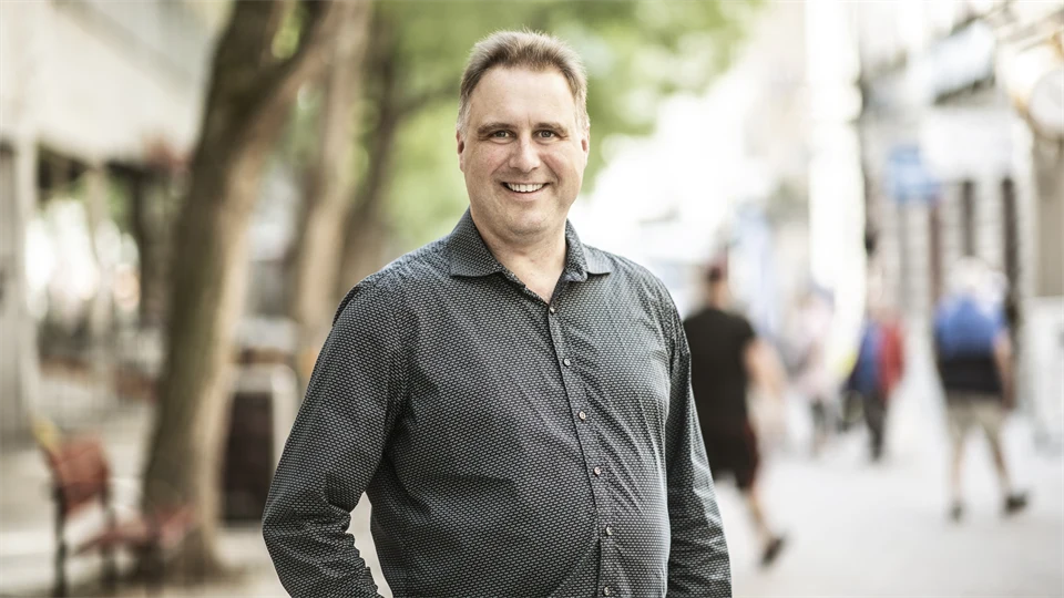 Man with light brown hair, wearing a gray shirt smiles at the camera