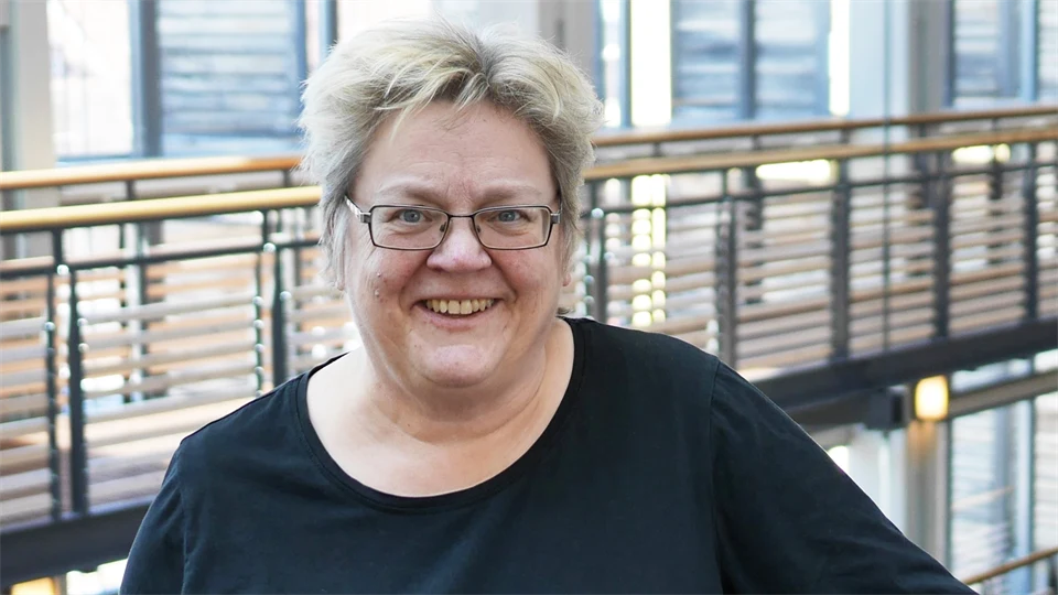 En kvinna står inomhus med fönster och ett räcke bakom sid. Hon har glasögon och en svart tröja. Hon ler. 