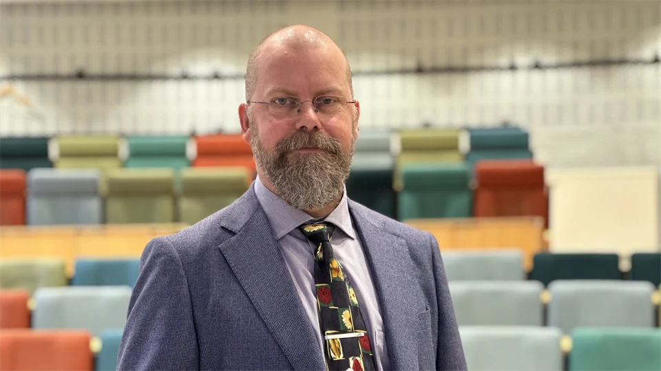 En man i blå kavaj visar upp sin doktorsavhandling i en sal med fägglada stolar