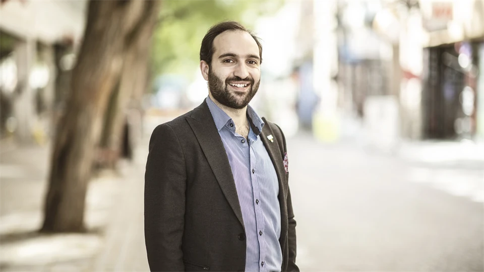 A man with dark brown hair, dark brown jacket and striped shirt smiles at the camera