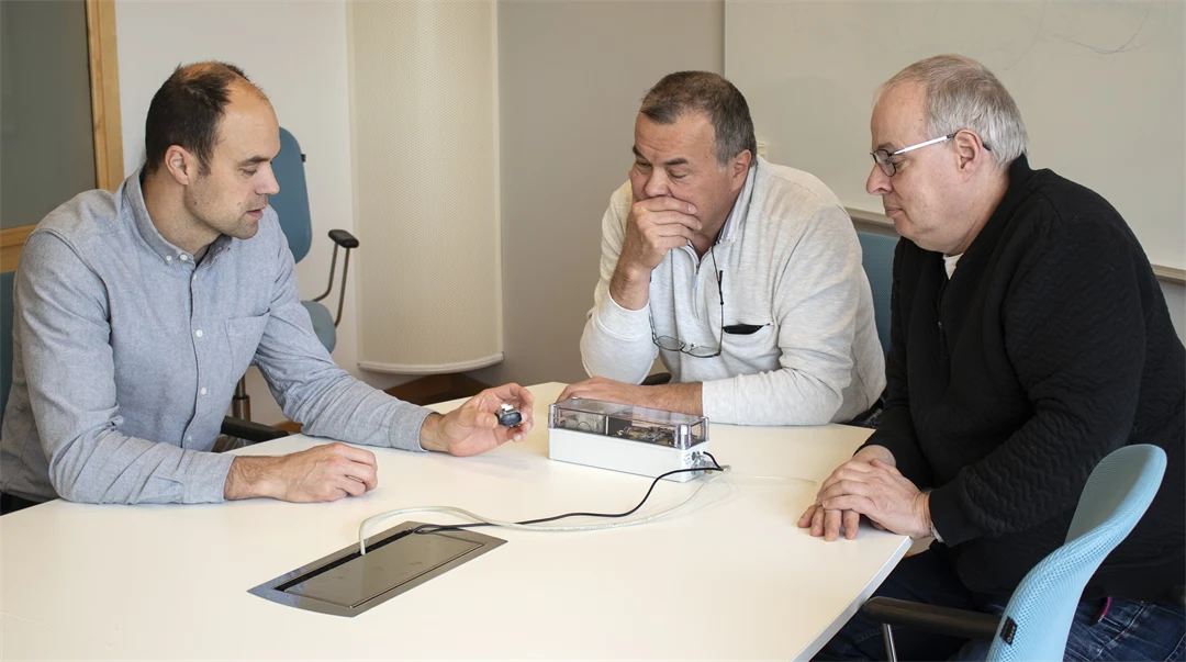 Three men sit at a table and discuss a prototype lying on the table.