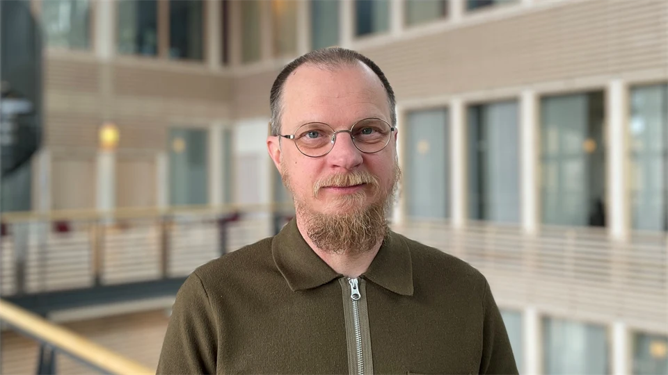 EN man med skägg och glasögon står i en ljushall. Bakgrunden är suddig.