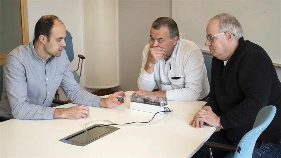 Three men sit at a table and discuss a prototype lying on the table.