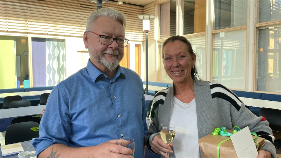 En mörkhårig kvinna i grå kofta står bredvid en gråhårig man i blå skjorta. Båda har champagneglas i händerna och ser glada ut. Kvinnan håller i ett paket.