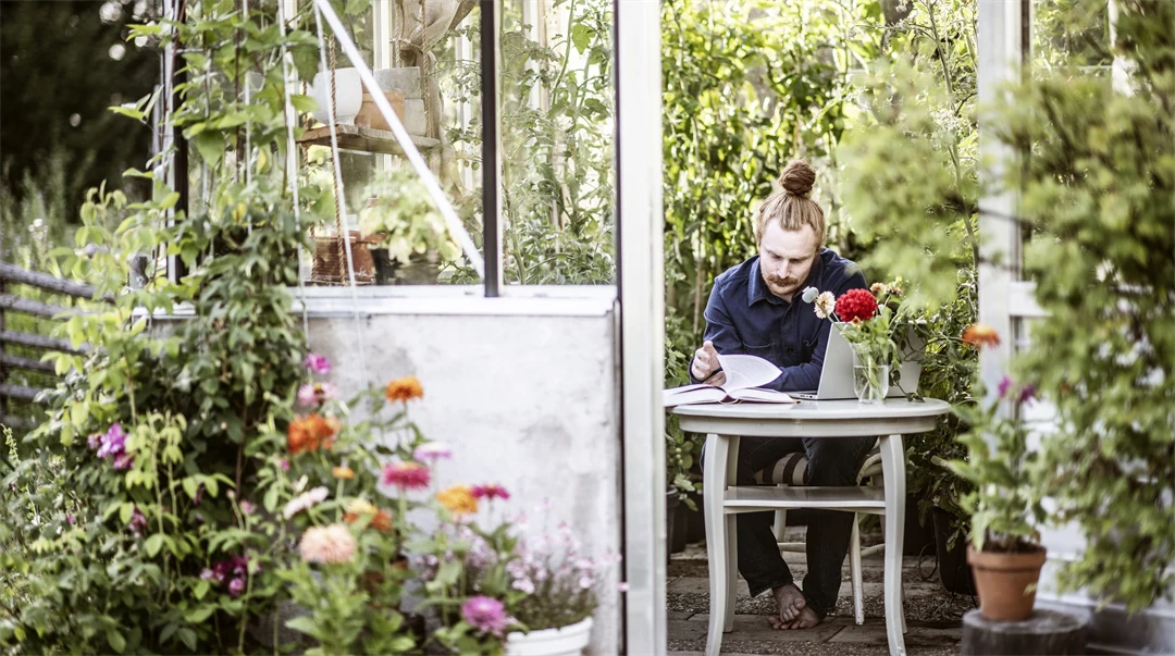 Manlig student sitter i  växthus och pluggar med laptop och böcker. 