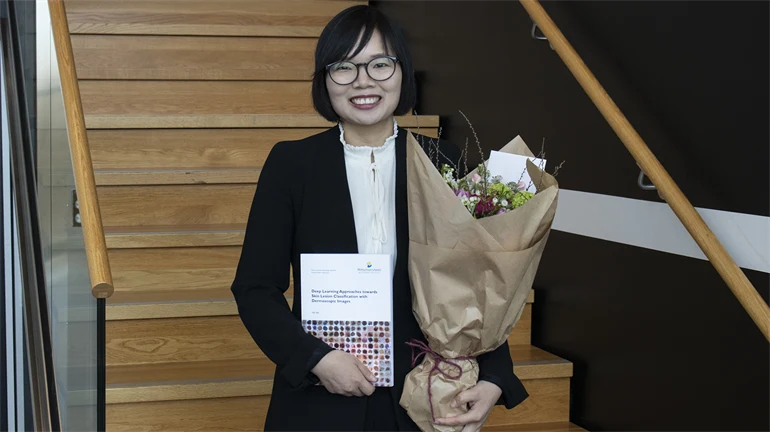 Yali Nie poses in front of the camera with her thesis and a bouquet of flowers.