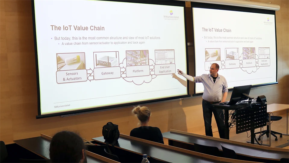 A man in a light blue shirt stands in front of a large screen and gives a presentation.