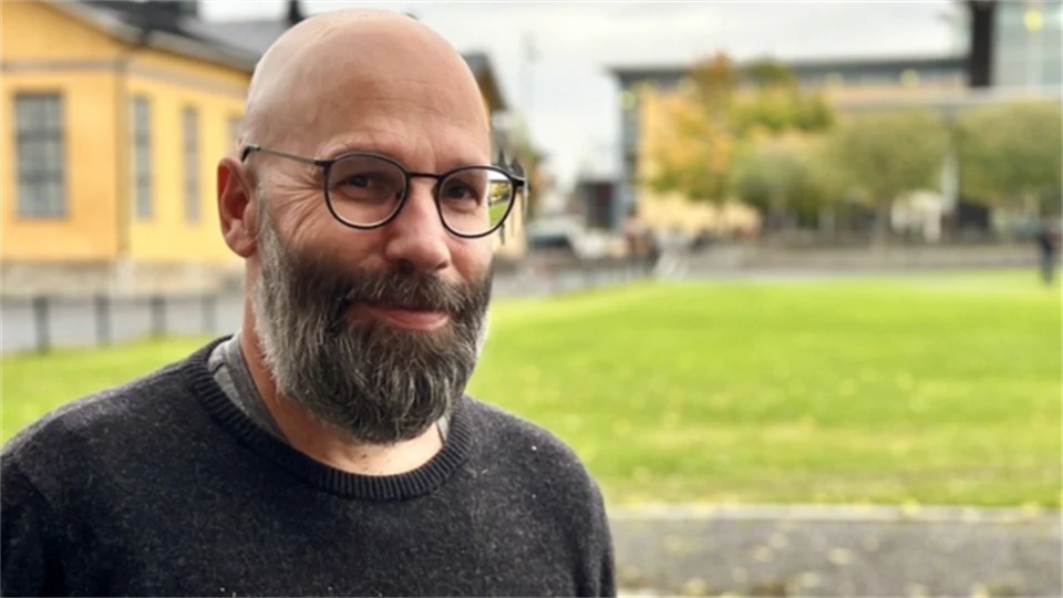 En man med glasögon och en mörkgrå tröja som står utanför framför gula byggnader och ler in i kameran