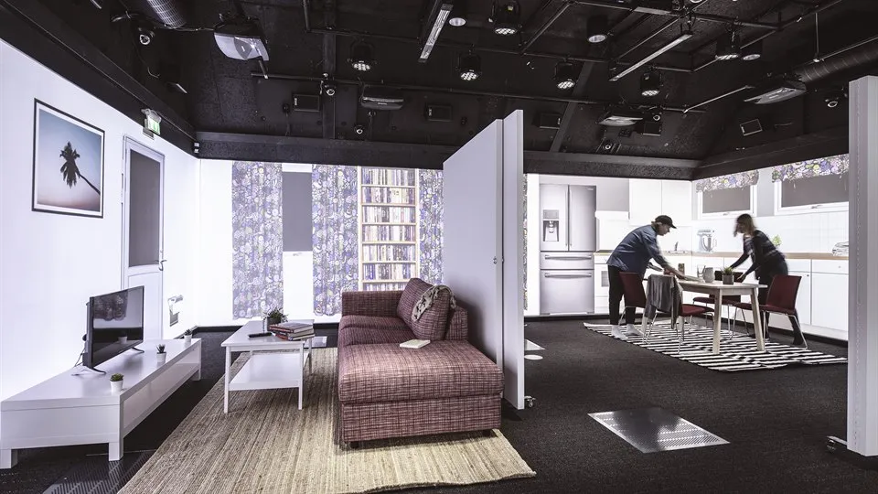 A large room with black floor and ceiling. The room looks like an apartment with partitions, furniture, and projections on the walls. Two people are standing at the kitchen table. There are lots of technical equipment in the ceiling.