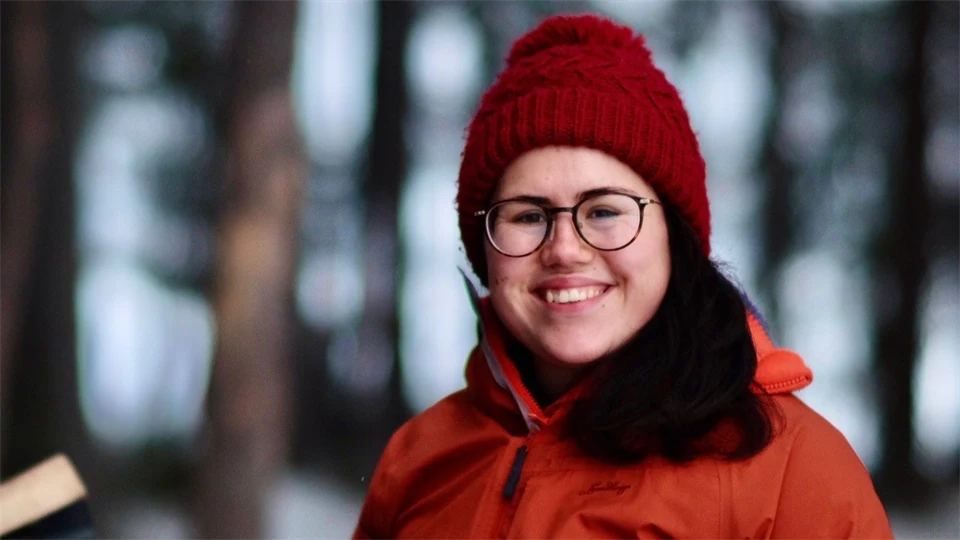 A woman with a red hat and dark brown hair wearing an orange winter jacket smiles at the camera. 