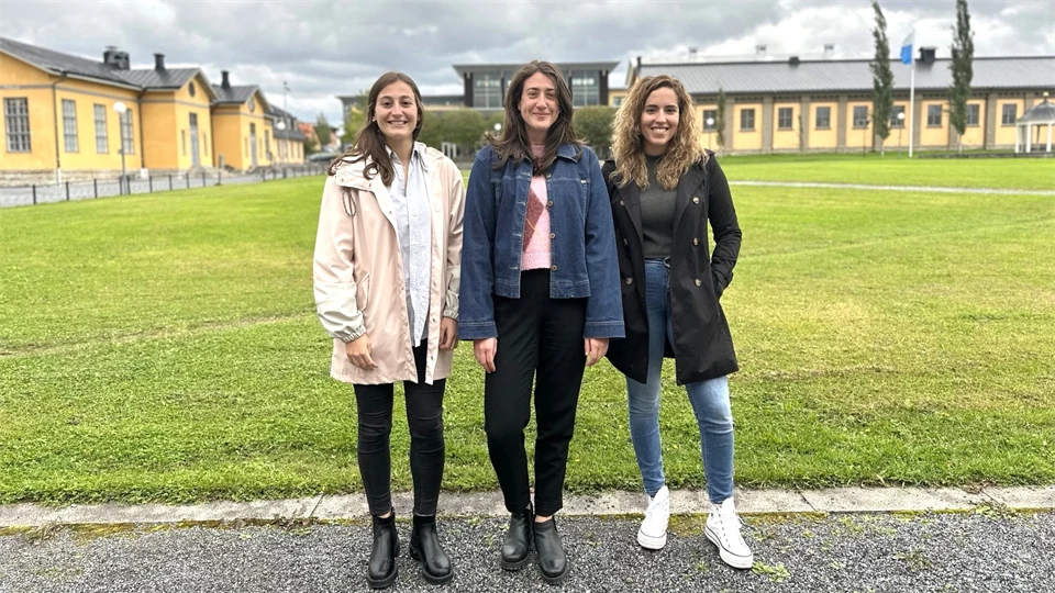 Tre unga kvinnor i ytterkläder står framför en stor gräsyta med hus i bakgrunden. De ler in i kameran. 