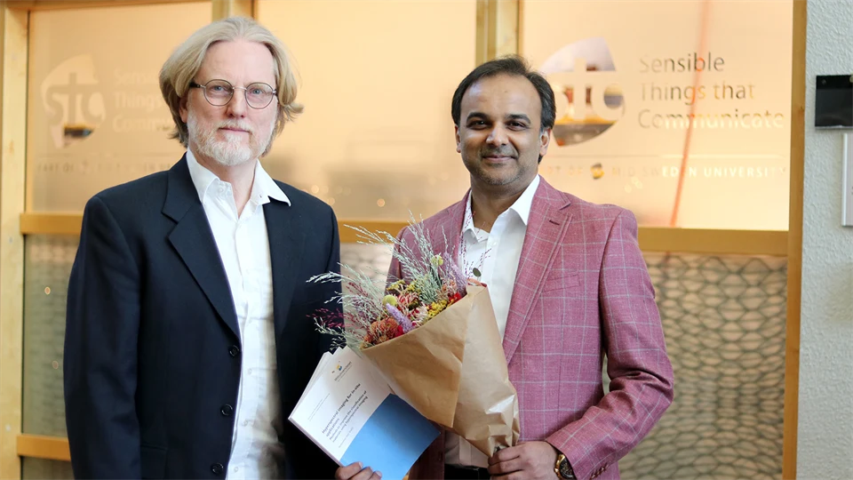 Two men stand side by side. One man is holding a bouquet of flowers and a doctoral thesis.