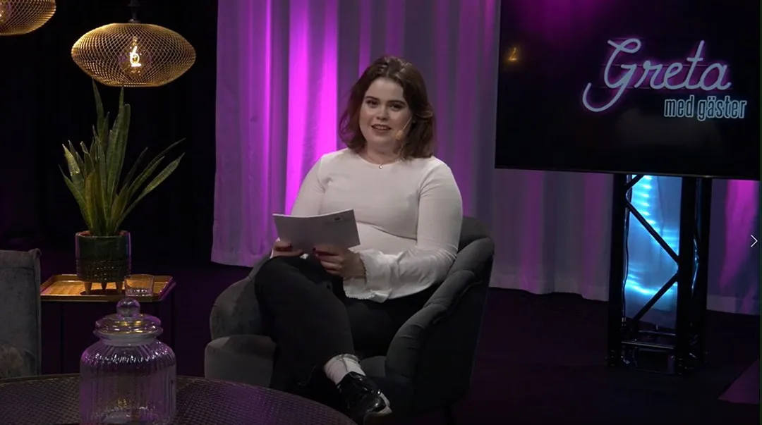 En kvinna sitter i en fåtölj i en studio, hon tittar in i kameran.