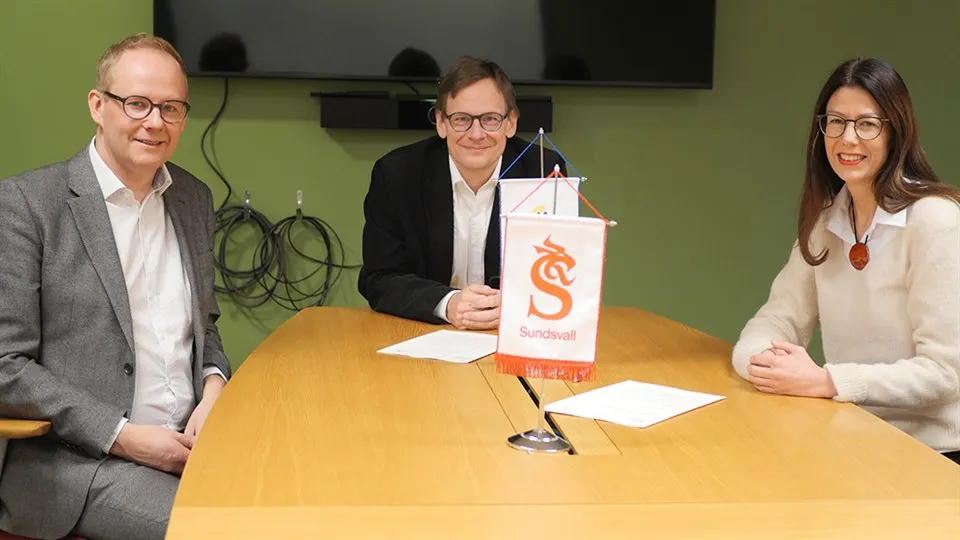 Två män och en kvinna sitter vid ett konferensbord med papper liggande framför sig.