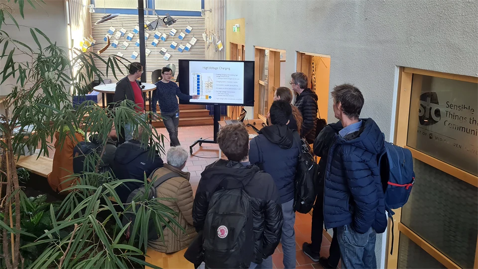 A group of people standing with their backs to the camera watching a television and a man presenting.