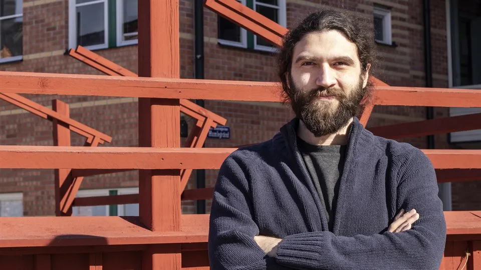 A man with dark hair and beard standing in front of a red fence.
