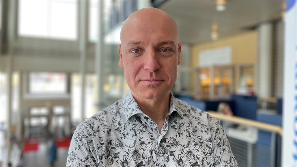 En man i mönstrad skjorta tittar in i kameran. Bakgrunden är suddig. 