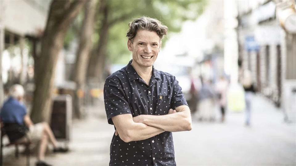 Man with brown hair, wearing a short-sleeved patterned shirt smiles at the camera