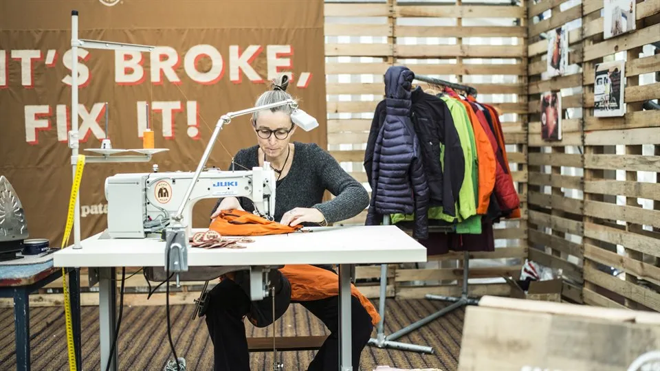 Woman sits at a desk and sews/repairs used clothes on a sewing machine