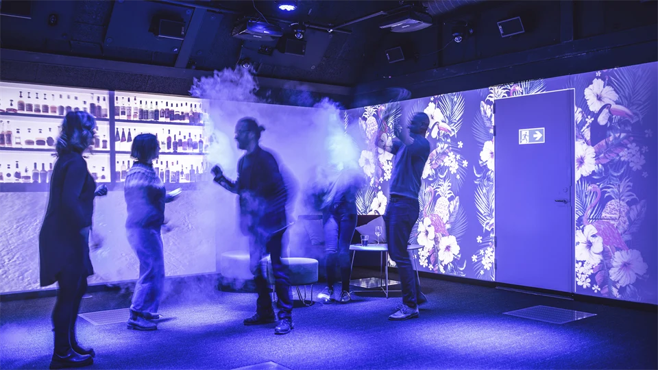 Fyra personer dansar i ett rum med dämpad belysning. På väggarna projiceras en nattklubbsmiljö med bar och en nödutgång. I ett hörn står några sittpuffar och ett lågt bord.