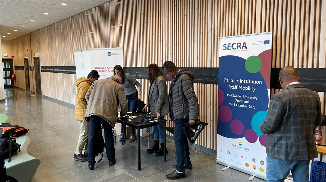 Några personer står vid ett bord. Bredvid bordet står en roll-up med texten "SECRA".