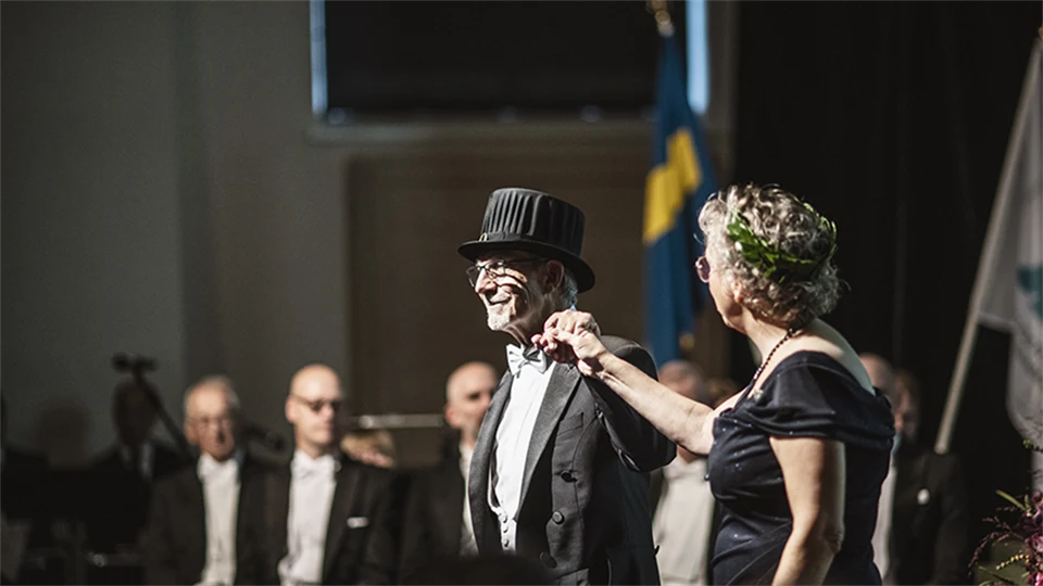 Two well-dressed people, one with a doctor's hat and one with a laurel wreath, stand next to each other on a stage and holding hands.