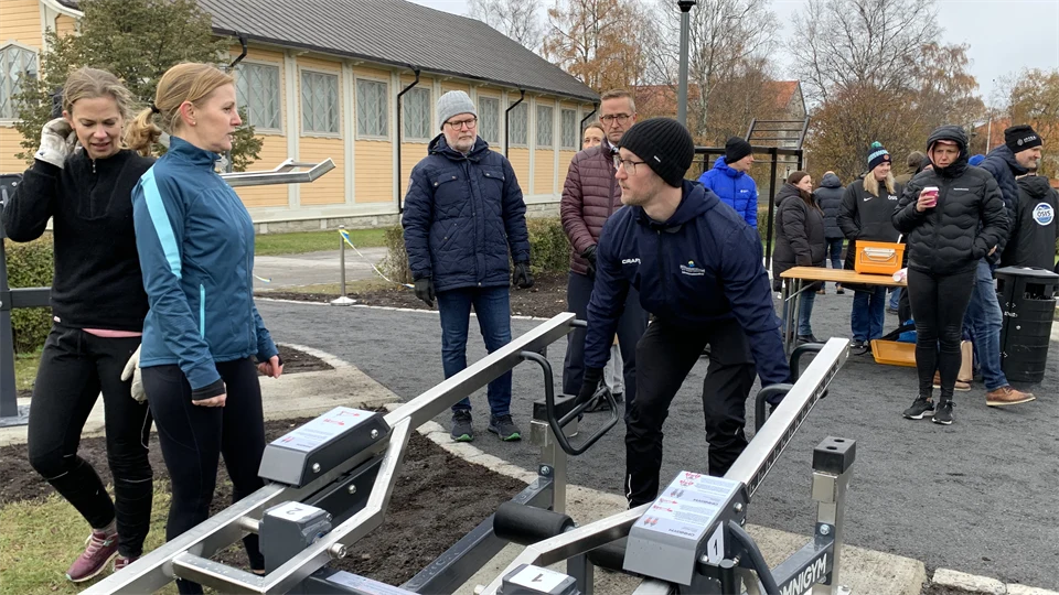 Människor står utomhus runt en träningsmaskin, andra står och betraktar i bakgrunden.