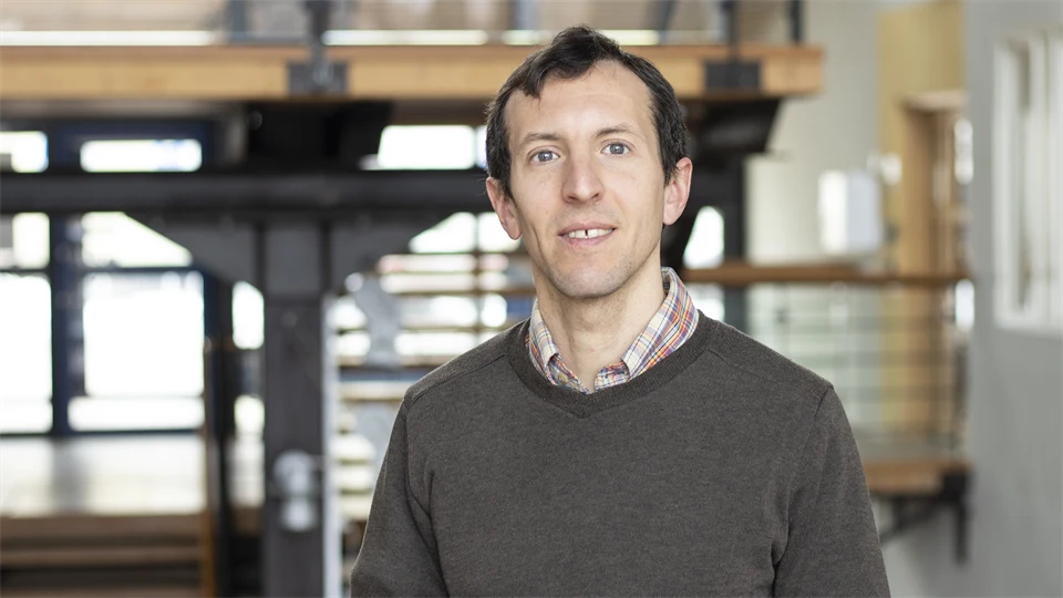 A man with dark hair, colorful shirt with a brown sweater over it.