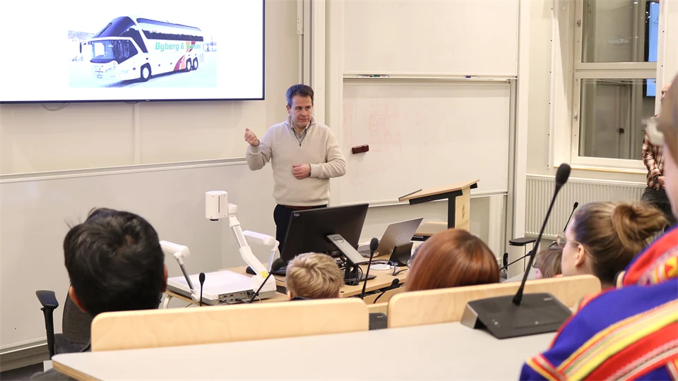 A man in a light shirt stands and presents to an audience