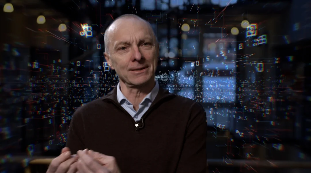 Man with light shirt and black knitted sweater in front of a digital background.