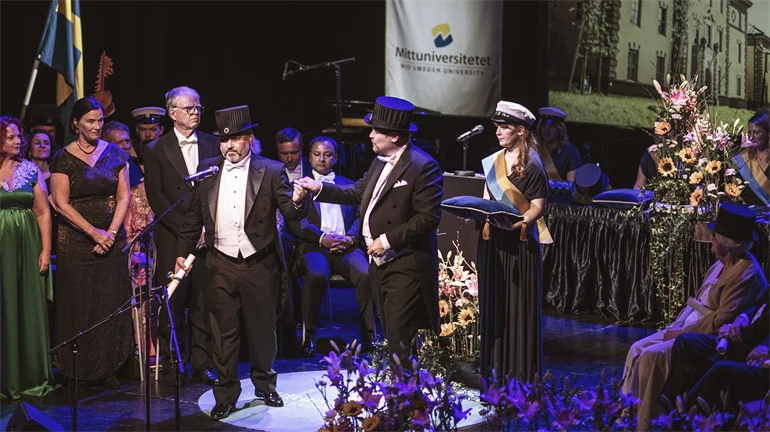 En finklädd person med doktorshatt står i rampljuset på en scen. 