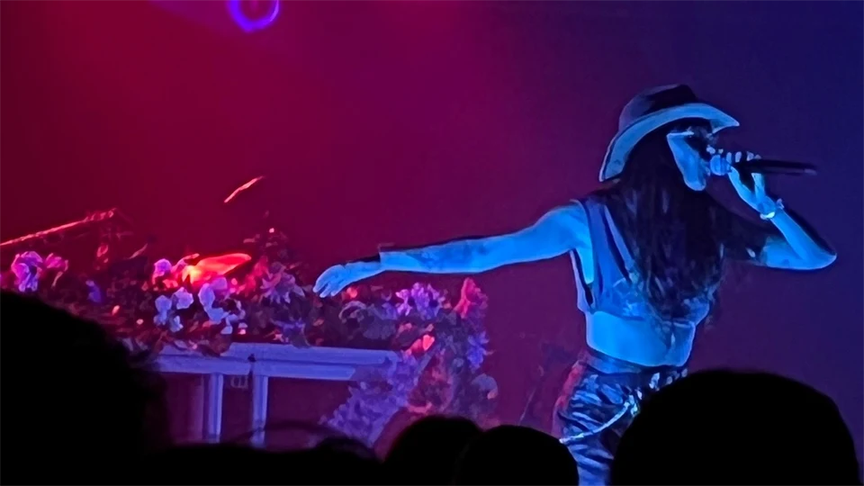 En kvinna står på en scen med mikrofon i handen. Hon sträcker ut ena armen. Ljuset är blått och rött.