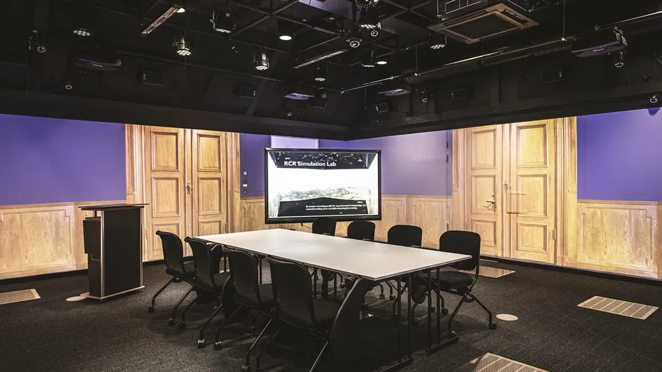 A large room with black floor and ceiling. Images from a grand meeting room are projected on the walls. The room is furnished with a large table and eight chairs, a computer screen and a lectern. There are lots of technical equipment in the ceiling.