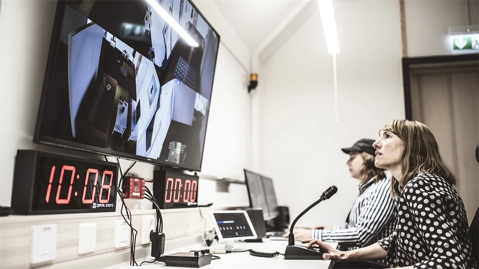 Två personer sitter vid ett långt arbetsbord. Framför dem finns flera datorskärmar, mikrofon och annan teknisk utrustning.