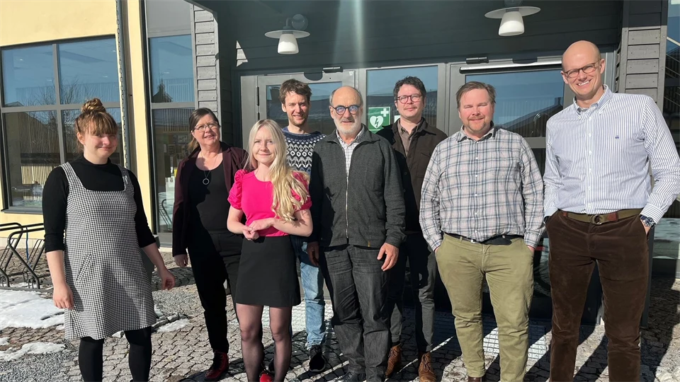 En grupp människor står framför entrén till en gul byggnad. Solen skiner.