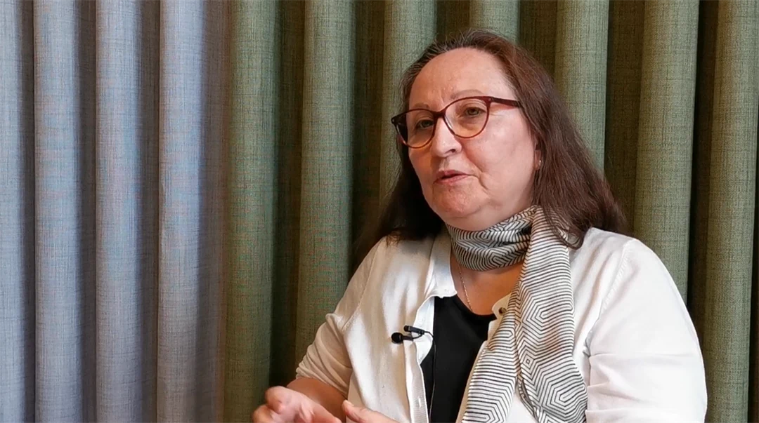 A woman with long brown hair wearing a white blouse and a green scarf is interviewed.