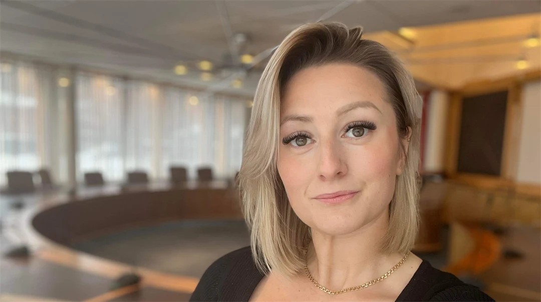 Kvinna med ljust och kort hår tittar in i kameran, kvinnan bär en svart stickad tröja och står framför ett stort mötesbord.