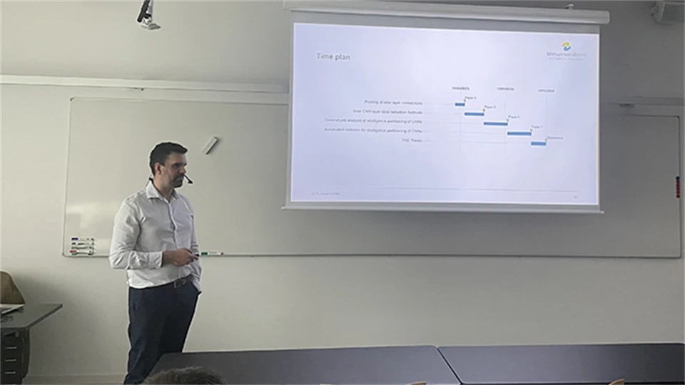 A man in a white shirt stands in front of a large screen and presents.