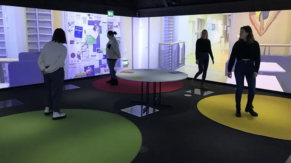Fyra personer står i ett rum. På golvet finns runda mattor i olika färger. Några av personerna står på mattorna. På väggarna projiceras bilder från en korridor i en skolbyggnad.