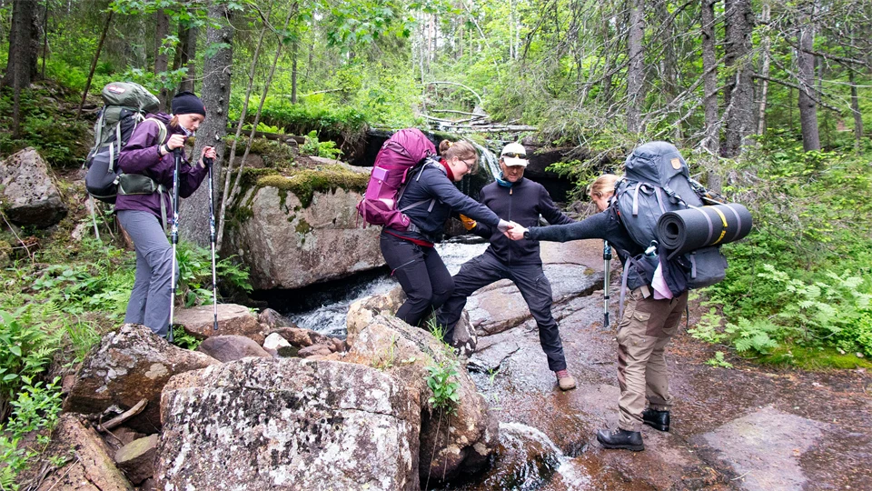 Skogsvandrare med ryggsäckar hjälps åt att passera en bäck.