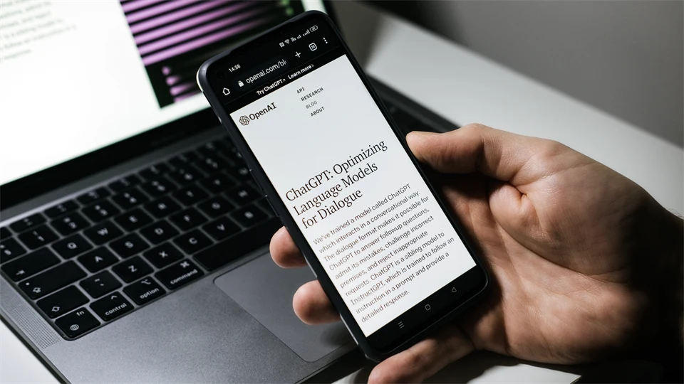 Hand holding a mobile phone with the text ChatGPT a computer in the background.