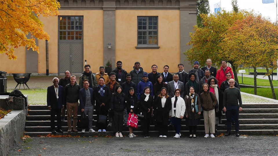 En grupp människor står på en stentrappa utomhus. I bakgrunden syns ett gult stenhus och träd med höstlöv.