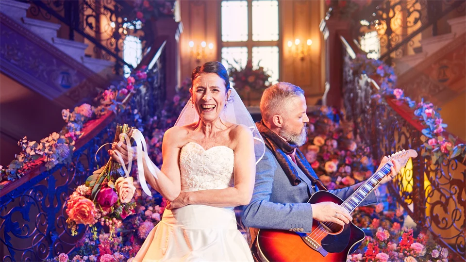 Kvinna i brudklänning, man i grå kostym med gitarr. Båda står framför en trappa fylld med blommor.