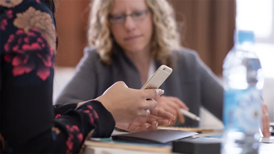 Female hand at a table holding a mobile phone, blurred woman in the background.