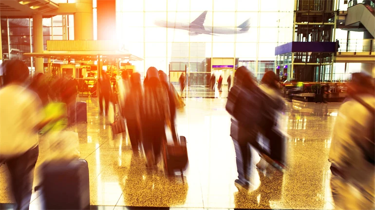 Människor går inomhus på flygplats med resväskor och ett flygplan syns genom fönstret utanför 